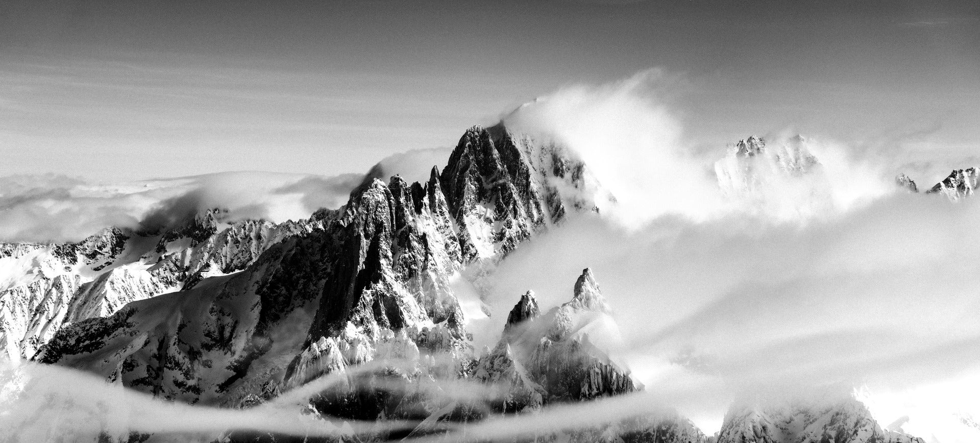 Black and white panoramic photograph of snow-covered mountain peaks rising above a sea of clouds.